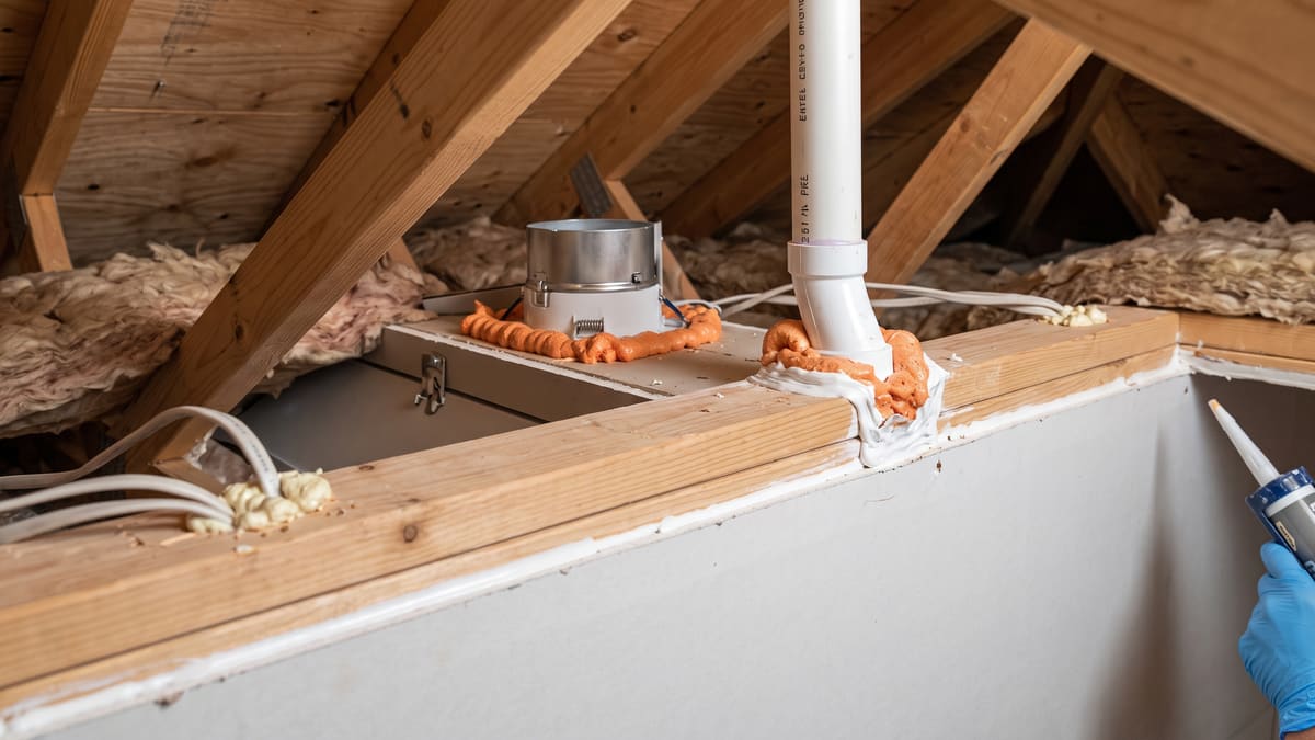 Orange spray foam and white sealant applied around a recessed light housing in an attic. Sealant also covers a PVC vent penetration and wiring at the top plate. A gloved hand holds a caulk gun.