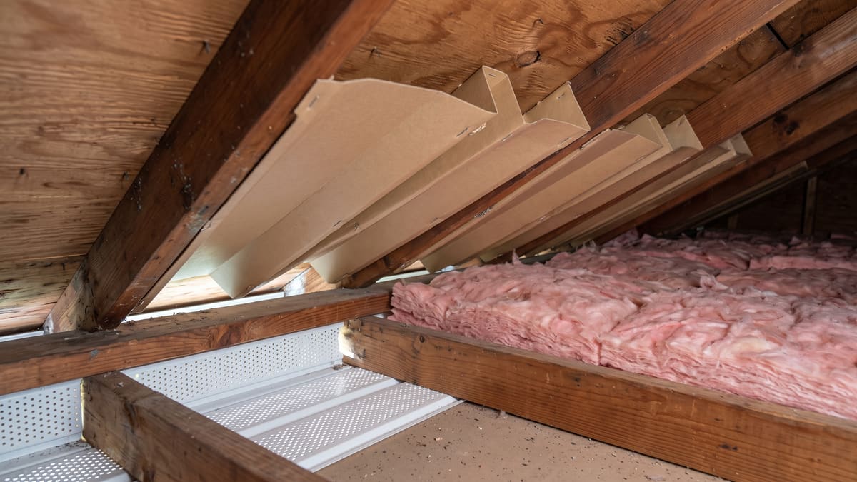 Cardboard baffles stapled between rafters at the eave, holding the airway open above the perforated vented soffit. Batt insulation lies alongside.