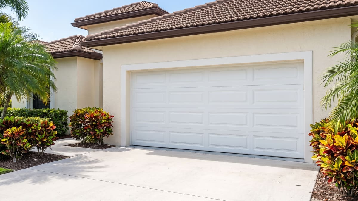 Closed white steel double-bay garage door running the full width of the opening. It has four sections of raised panels. The cream stucco Florida home has a barrel tile roof and a wide concrete driveway.