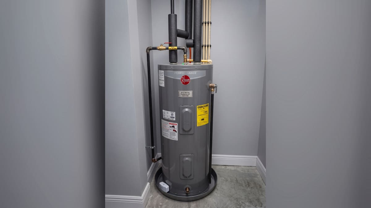 A grey Rheem tank water heater standing in a drain pan in a utility closet. Insulated supply lines and a shut-off valve run overhead.
