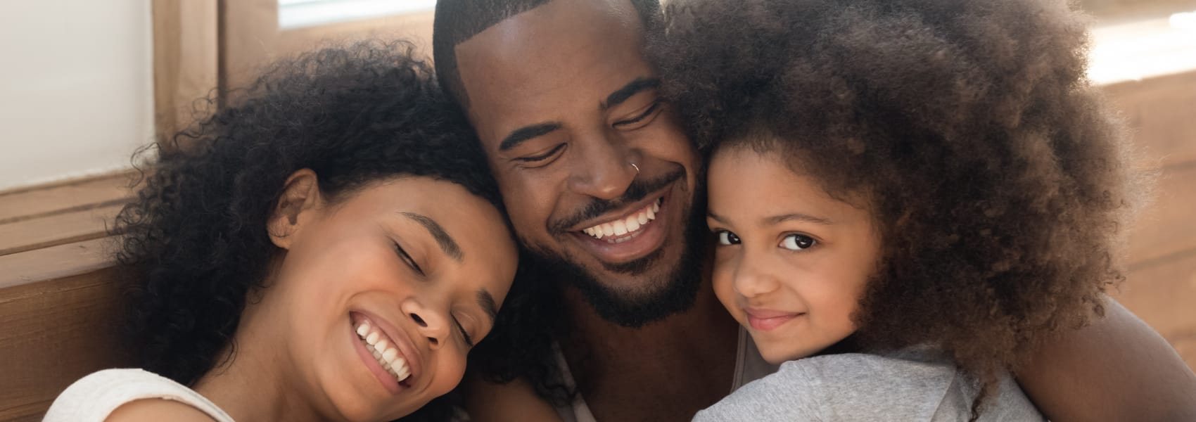 A mother, father, and daughter smiling closely together in front of a wood-paneled wall, the mother and father with eyes closed and the daughter looking at the camera