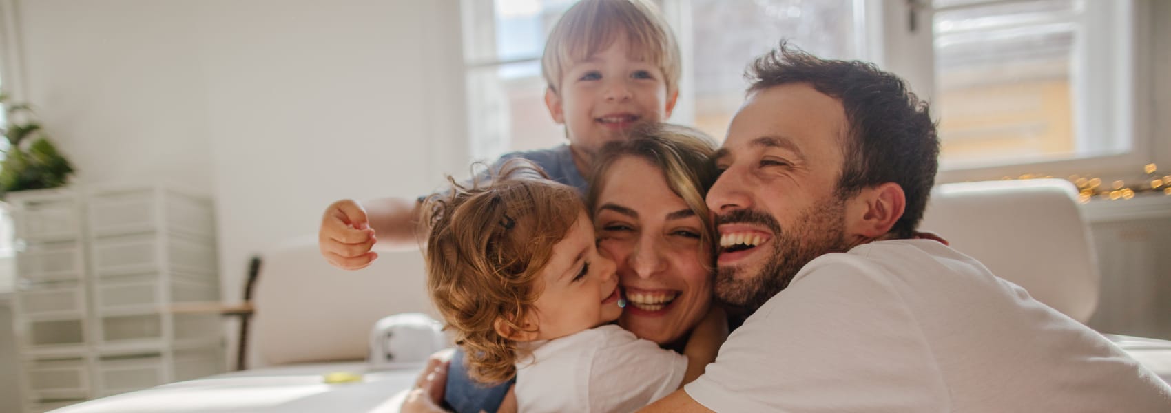 A mother and father embracing with a toddler girl kissing the mother’s cheek and a young boy behind them raising his arm, all smiling together in a bright room with tall windows