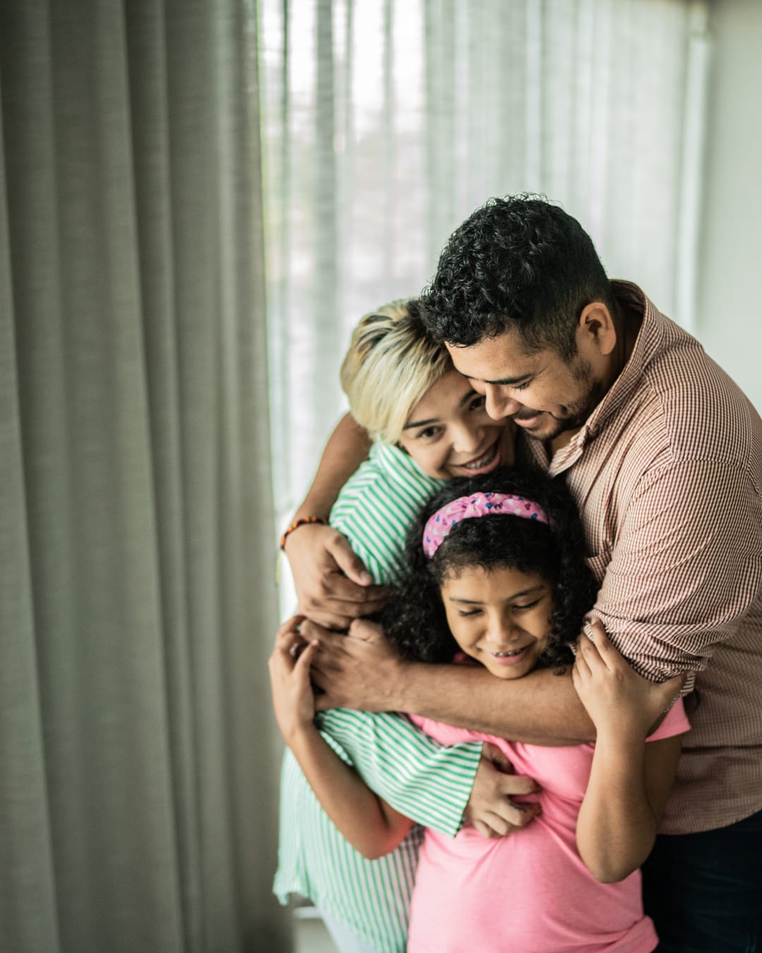 Un padre, una madre y su hija abrazados en casa junto a una ventana con cortinas finas, todos sonriendo