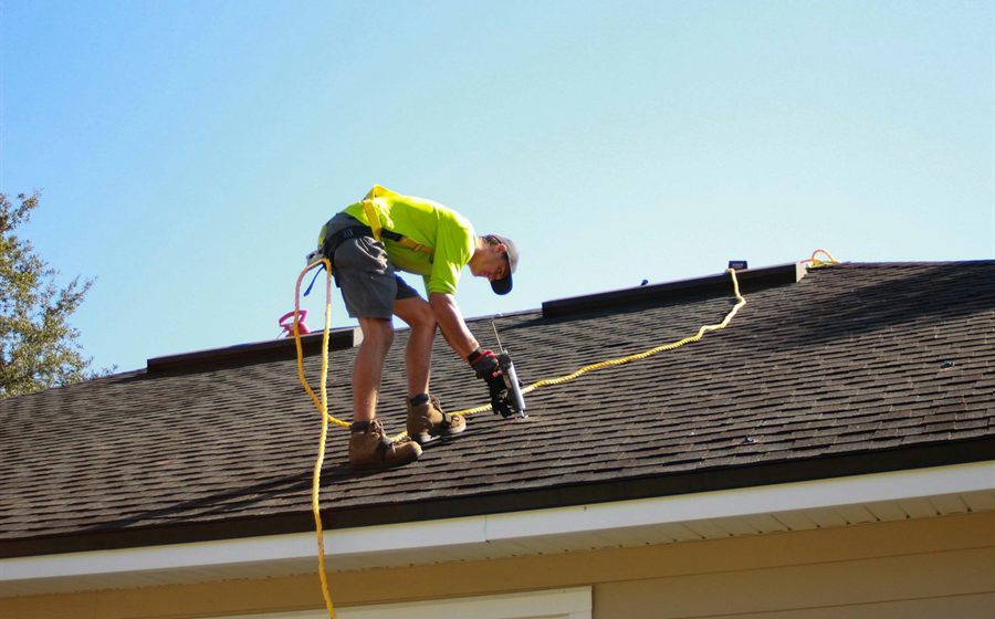 Roofer in a safety harness fastening shingles on a single-story home